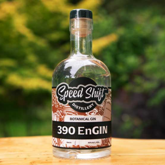 Clear glass bottle filled with Speed Shift Distillery's 390 EnGin botanical gin wrapped in a mocha brown label sits on a wooden table.
