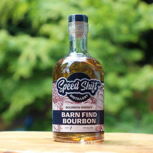 Clear glass bottle filled with Speed Shift Distillery's Barn Find Bourbon Whisky wrapped in a nut brown colored label sits on a wooden table.