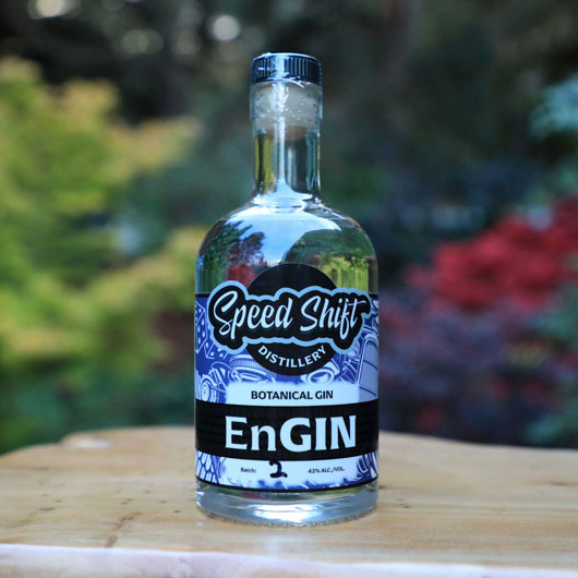 Speed Shift Distillery's Engin Botanical Gin Clear bottle with deep purply-blue and white label featuring motor parts, sitting on a live edge wood slab outside on a sunny day.