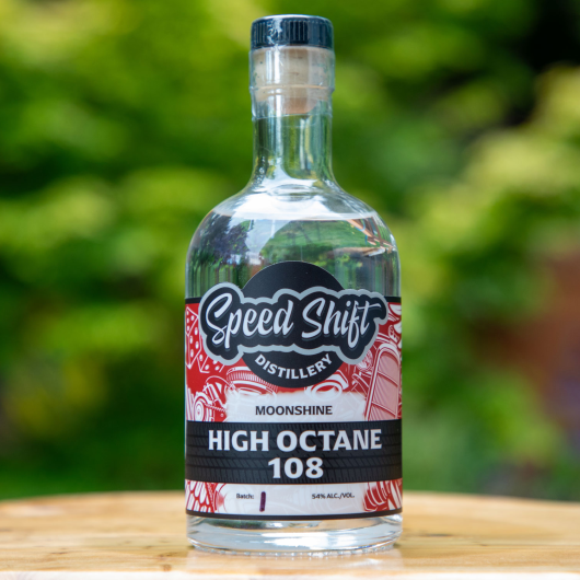 Clear glass bottle containing Speed Shift Distillery's High Octane 108 Moonshine sits on a wooden table.