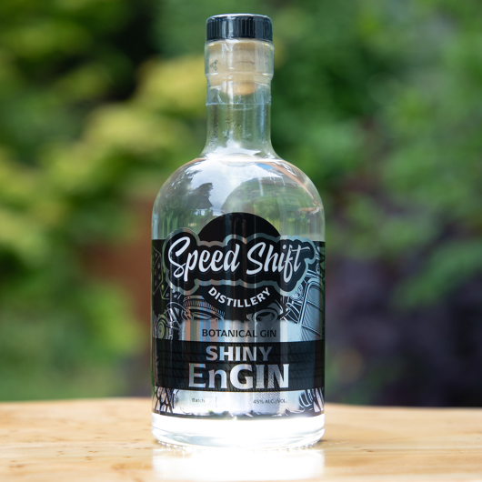 Clear glass bottle filled with Speed Shift Distillery's Shiny EnGin botanical gin wrapped in a shiny, chrome label sits on a wooden table.