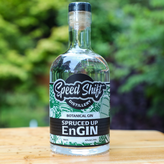 Clear glass bottle of Speed Shift Distillery's Spruced Up EnGin botanical gin wrapped in a deep green label sits on a wooden table.